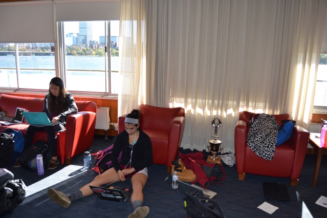 Teammates Michelle Lauer (left) and Sylvia Sarnik sit in the boathouse in the hours before making MIT history. The space has a camped-out family feel. Nervous first-year students lob earnest question at their elders: “What should I eat before the race?” (Photo: Sam Hunter Magee/Arts at MIT)