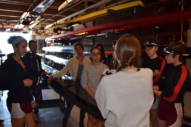 Coxswain Chloe Thacker (center right, with back turned) briefs members of the Varsity 8 about the high wind conditions along the 5-km course. Left to right: Sylvia Sarnik, Claire Martin-Doyle (head coach), Priya Veeraraghavan (captain), Sharon Wu (captain), Kelly Barton, Valerie Hunter, Elizabeth Martin, and Annika Rollok. “Wear layers,” says Thacker. “Be ready for water in the boat.” (Photo: Sam Hunter Magee/Arts at MIT)